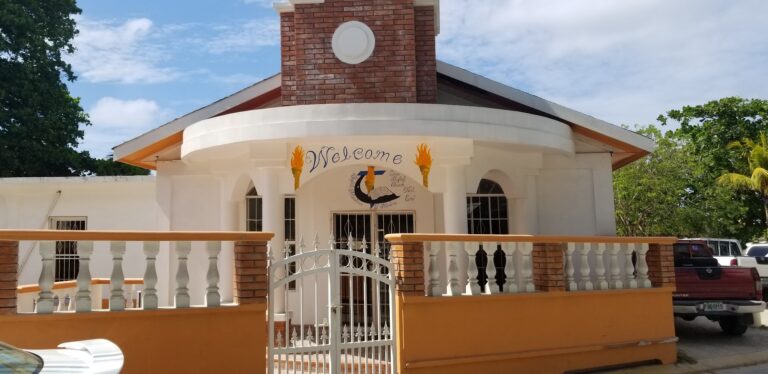 A white church with a brick steeple, decorative columns, and a "Welcome" sign featuring a cross, flames, and a Bible, located in Roatan, Honduras.