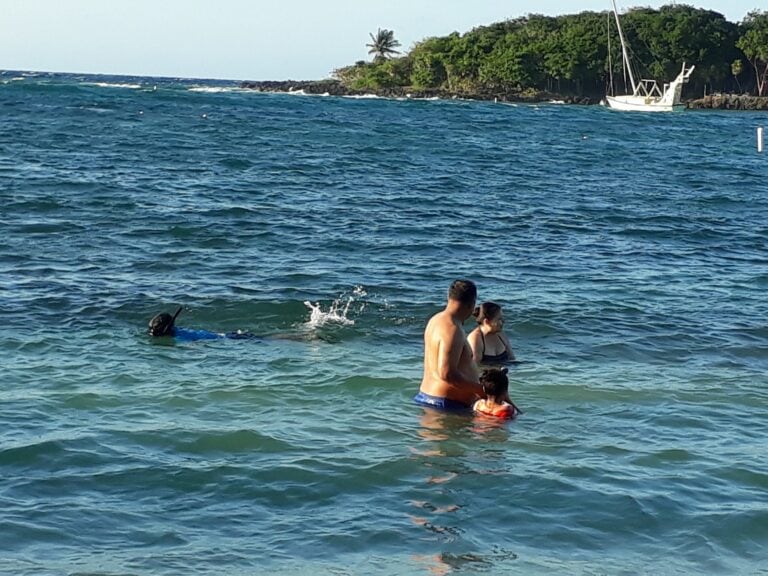 People swimming and relaxing in the ocean near a rocky, green coastline with a sailboat anchored off Roatan, Honduras.