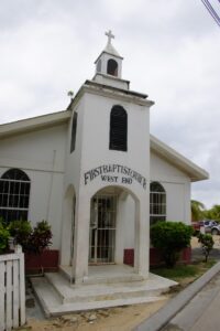 The white First Baptist Church building with a small steeple and cross on top, located in West End, Roatan, Honduras, is shown with black window shutters, decorative iron bars on the entrance door, and surrounded by greenery under a cloudy sky.