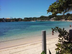 A sandy beach with clear turquoise water, boats anchored offshore, and lush green trees along the shoreline in Roatan, Honduras, visible under a bright blue sky.