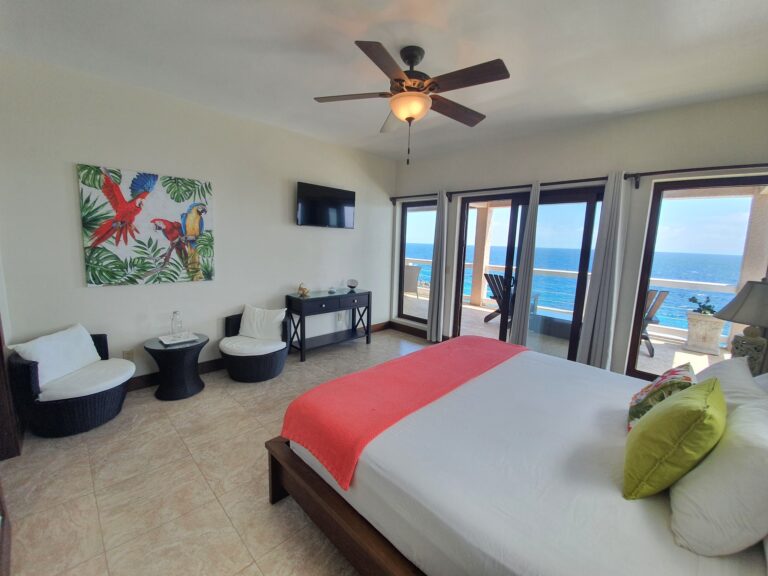 A bedroom in a Roatan hotel or vacation rental features a view of the ocean through glass sliding doors, with a wooden bed frame, colorful pillows, a ceiling fan, a small black table with chairs, a vibrant parrot and tropical leaf painting on the wall, and a balcony with outdoor seating overlooking the sea in Honduras.