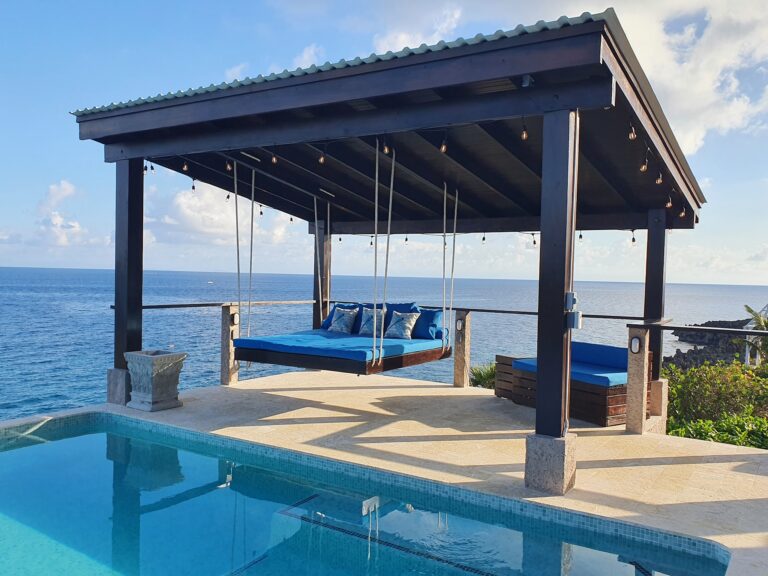 A shaded outdoor seating area with a swing in front of the ocean, overlooking Roatan in Honduras, featuring a decorative stone planter, string lights, and a view of the sea and sky.