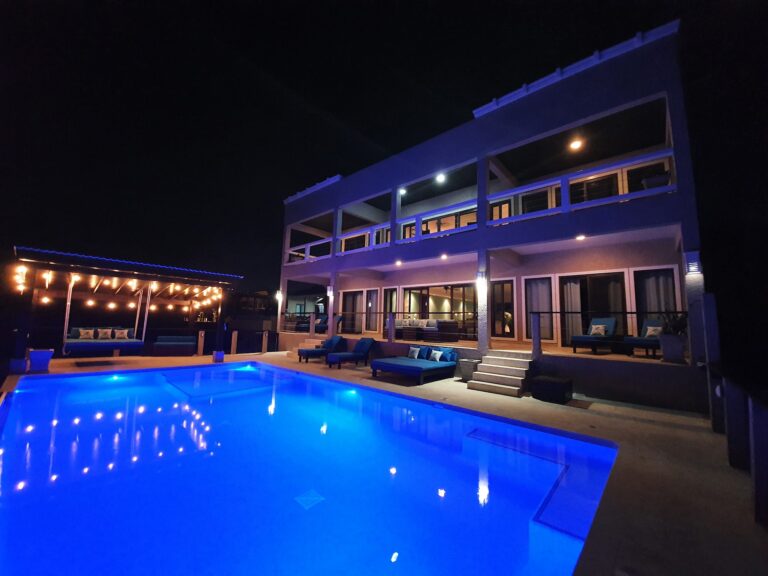 A modern multi-story building in Roatan, Honduras, features a spacious outdoor area with a swimming pool illuminated by blue lights, surrounded by seating and well-lit architectural details at night.