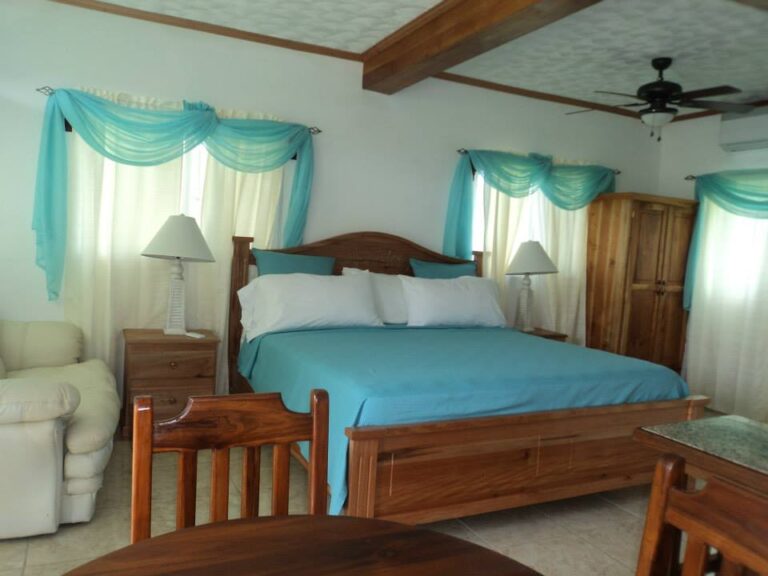 A cozy bedroom in Roatan, Honduras, features a wooden bed with a blue bedspread, matching curtains with light blue swags, two bedside lamps, a white armchair, a wooden wardrobe, and a ceiling fan, with natural light filtering through the windows.