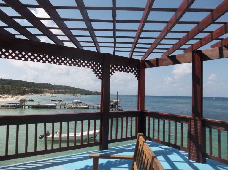 A view from a wooden balcony overlooking Roatan, Honduras, featuring boats anchored in the turquoise sea, a sandy beach with palm trees, and a partly cloudy sky.