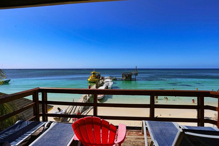A view from a beachfront balcony in Roatan, Honduras, overlooking the clear turquoise waters, sandy beach, a small pier with boats, and people swimming and relaxing in the ocean.