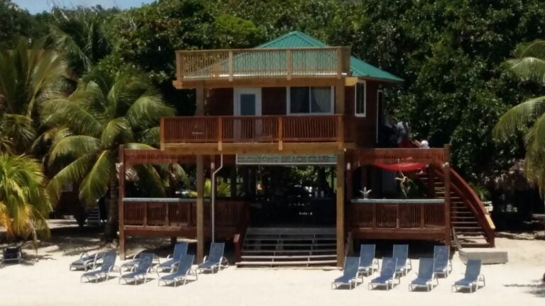 A multi-level wooden beach house with a green roof and outdoor deck seating is situated on the sandy shore of Roatan, Honduras, surrounded by lush palm trees and featuring several lounge chairs in front.
