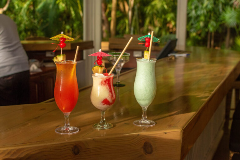 Three colorful tropical cocktails with fruit garnishes and paper umbrellas on a wooden bar counter in Roatan, Honduras, with lush jungle greenery in the background.