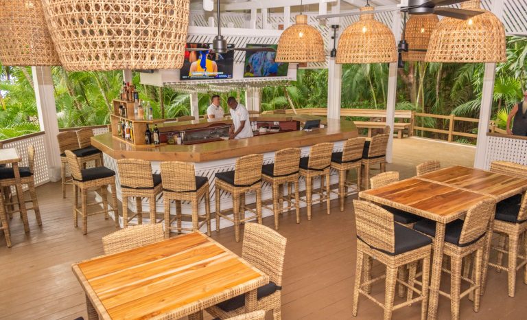 A tropical bar and restaurant in Roatan, Honduras, features wooden tables and wicker chairs, a curved wooden bar with bartenders, woven pendant lights, and lush greenery visible through open sides.
