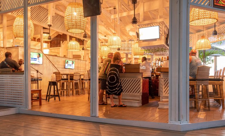 A group of people socializes at an indoor bar with wicker pendant lights and wooden decor, located on a patio overlooking palm trees in Roatan, Honduras.