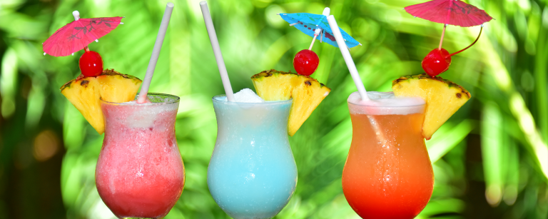 Three colorful tropical cocktails garnished with pineapple slices, maraschino cherries, and miniature umbrellas are displayed against a lush green background, representing a vibrant outdoor setting in Roatan, Honduras.