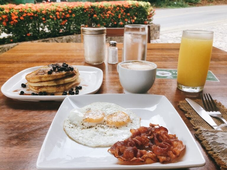 A breakfast spread featuring a stack of blueberry pancakes with syrup and berries, fried eggs, crispy bacon, and a variety of beverages including a glass of orange juice, a cup of coffee, and two glasses of water on a wooden table outdoors in Roatan, Honduras.