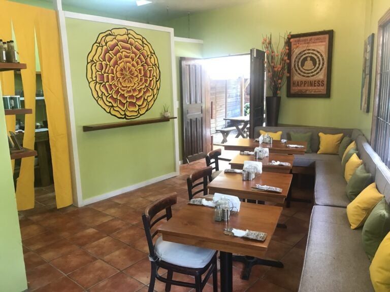 A cozy indoor dining area in Roatan, Honduras, features wooden tables with white napkins and glassware, a light green wall decorated with colorful artwork including a large yellow and red floral mural, and a seating corner with yellow pillows, while an open door reveals an outdoor patio with a wooden picnic table.