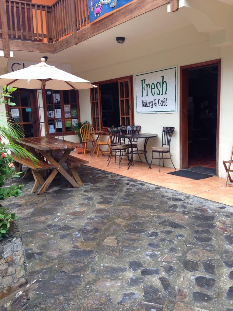 A storefront in Roatan, Honduras, features a sign for "Fresh Bakery & Café," with outdoor seating that includes wooden and metal tables and chairs, a patio umbrella, and lush greenery.