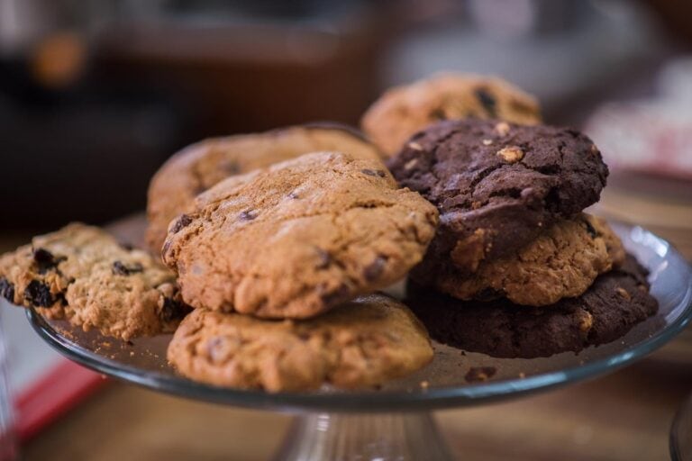 A glass cake stand holding a variety of freshly baked cookies, including chocolate chip and double chocolate cookies, is displayed on a wooden table in Roatan, Honduras.