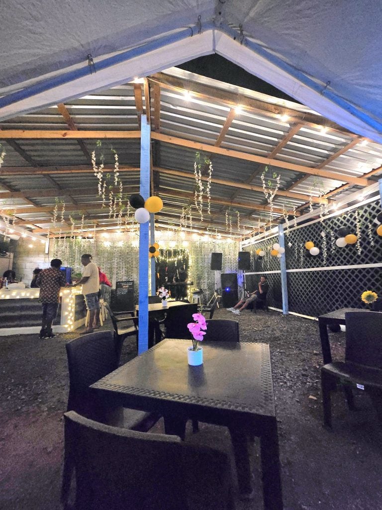 An outdoor event space in Roatan, Honduras, decorated with hanging string lights, balloons, and flowers, featuring a tented roof and people socializing near a table with a potted orchid, set against a lattice fence and evening sky.