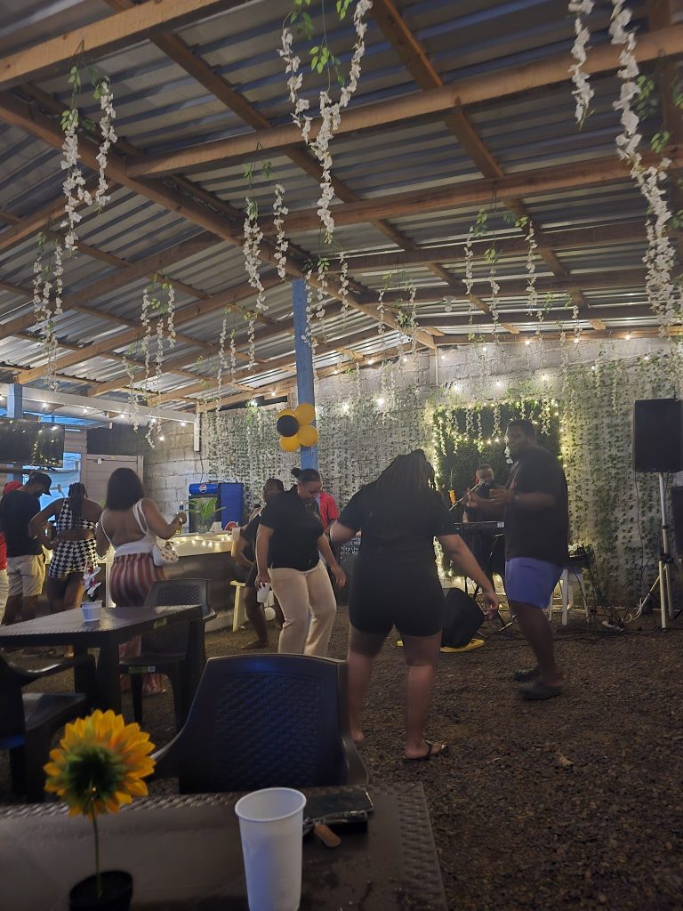 People are enjoying live music and socializing indoors at a rustic event space in Roatan, Honduras, decorated with hanging white flowers, string lights, and a brick wall backdrop.