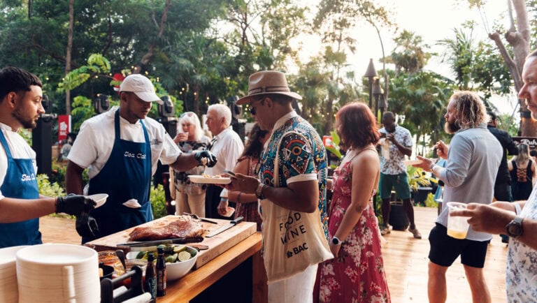 Crowd of people enjoying a barbecue event outdoors in Roatan, Honduras, with chefs preparing grilled meat and tropical greenery in the background.