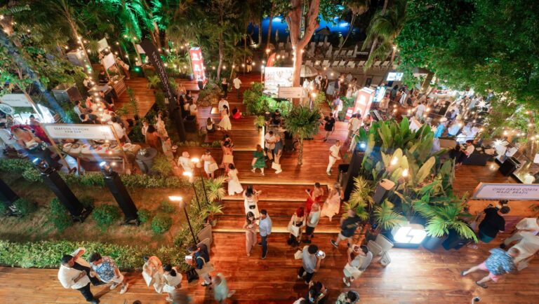 An aerial view of a lively outdoor night market or event in Roatan, Honduras, featuring wooden flooring, lush greenery, string lights, food stalls, and many visitors mingling under the trees