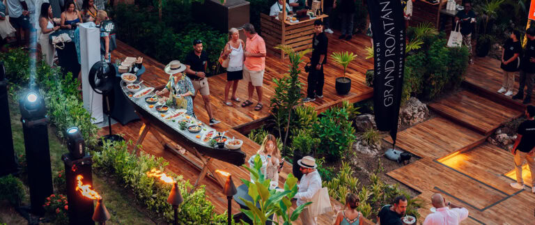 Guests enjoy a buffet-style dinner event on a wooden deck surrounded by lush greenery in Roatan, Honduras, with a catering table filled with various dishes and chefs serving food.