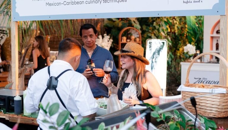 Three people engaging in a culinary tasting at a food stall in Roatan, Honduras, with tropical foliage and a decorated booth in the background.