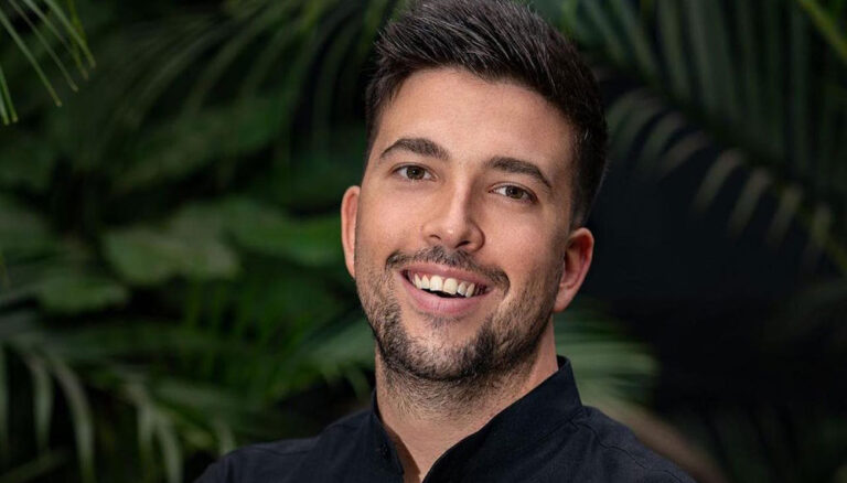 A smiling young man with short dark hair and a beard stands outdoors against a background of lush green tropical plants in Roatan, Honduras.