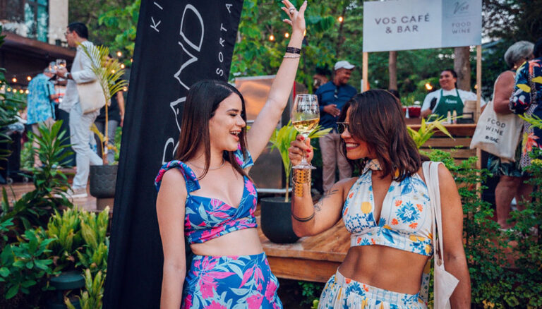 Two women celebrating with glasses of white wine at an outdoor event in Roatan, Honduras, surrounded by tropical plants and string lights