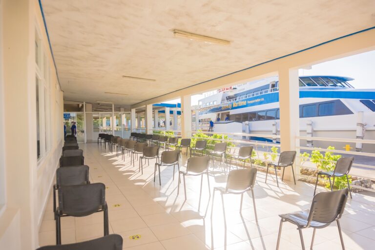 A seating area with black chairs and large windows overlooking a ferry docked at Roatan, Honduras, under a beige ceiling with fluorescent lights.