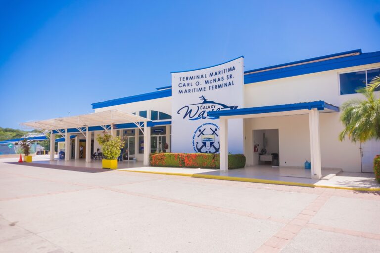 A white and blue maritime terminal building with signs indicating "Roatan" and "Honduras," featuring a covered waiting area, palm trees, and a clear blue sky in Roatan, Bay Islands, Honduras.