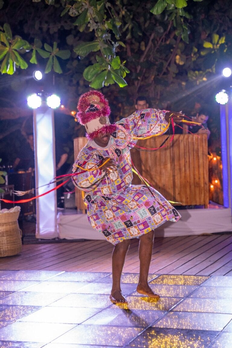 A person performing a traditional dance wearing a colorful, patchwork-style costume and a decorated mask, at an outdoor nighttime event on Roatan in the Bay Islands of Honduras, with a DJ booth, lighting, and lush green foliage in the background.