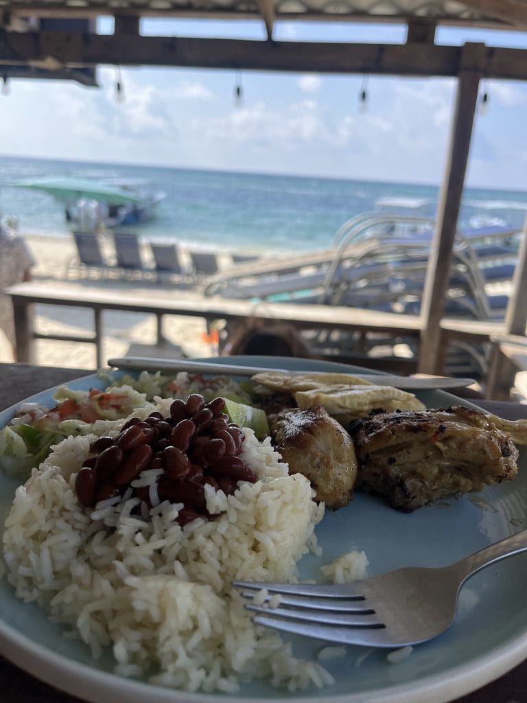 A plate of food including rice, beans, grilled chicken, and vegetables is served outdoors under a roof on a beach in Roatan, Honduras, with lounge chairs, a boat, and the ocean visible in the background.