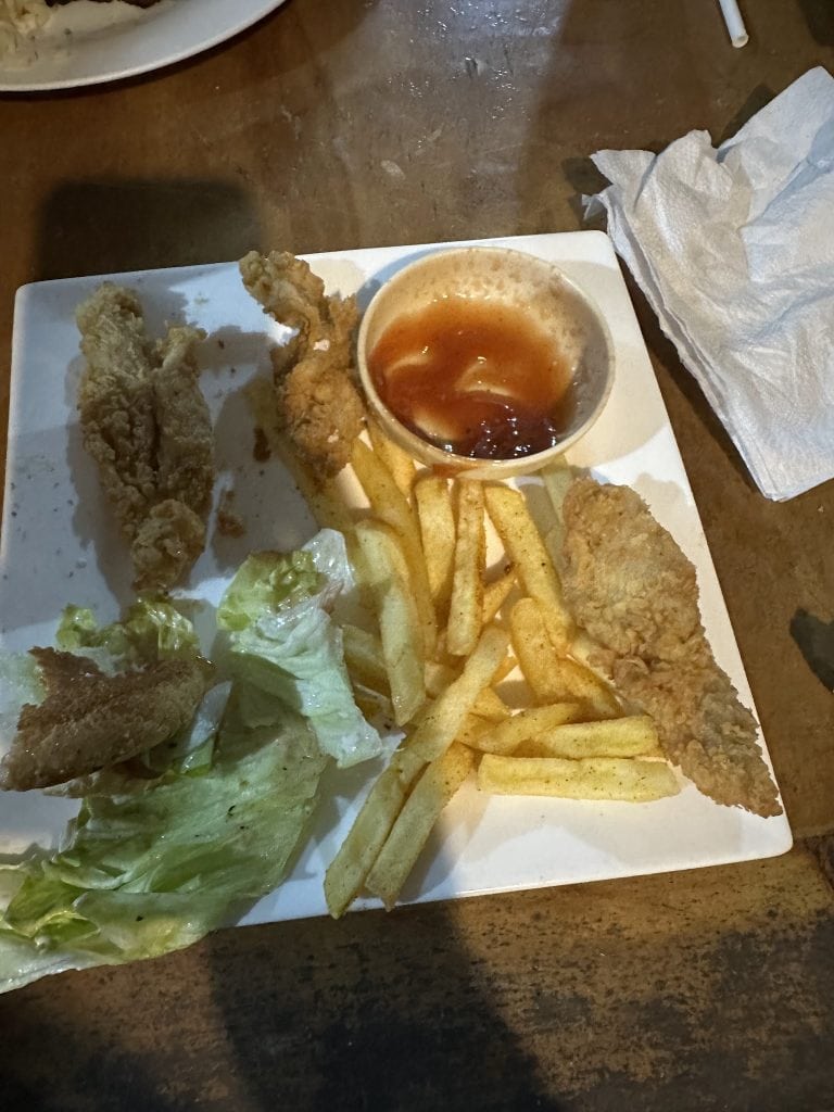 A plate of fried chicken, French fries, salad, and a cup of dipping sauce is displayed on a wooden table in Roatan, Honduras.