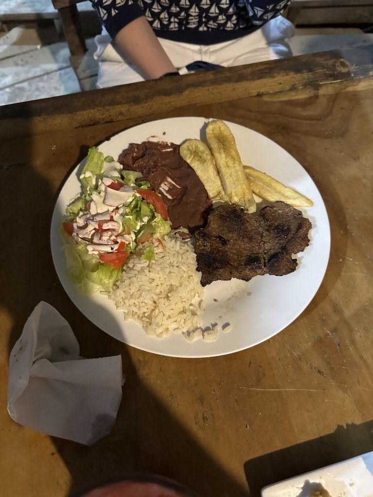 A plate of Honduran cuisine served on a wooden table in Roatan, Honduras, features rice, grilled meat, fried plantains, a salad with lettuce, tomatoes, radishes, and shredded cheese, with part of a person's arm and attire visible in the background.