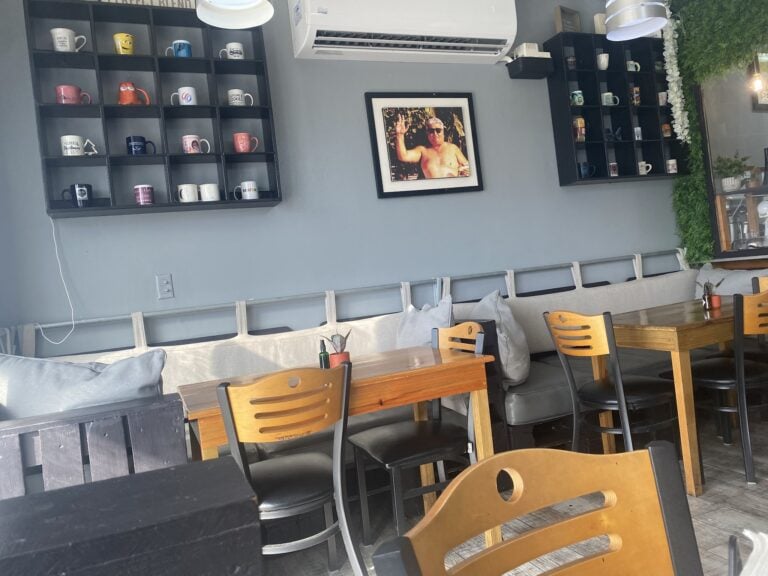 A cozy indoor seating area in a Roatan, Honduras café features wooden chairs, a long cushioned bench with pillows, black shelving displaying colorful coffee mugs, and a framed picture of a shirtless man waving, with greenery and decorative white flowers along the wall.