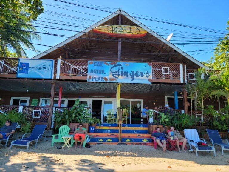 A group of people relax on lounge chairs on the sandy beach in front of a two-story restaurant called Ginger's in Roatan, Honduras, with colorful signage and lush green tropical plants surrounding the outdoor dining area.
