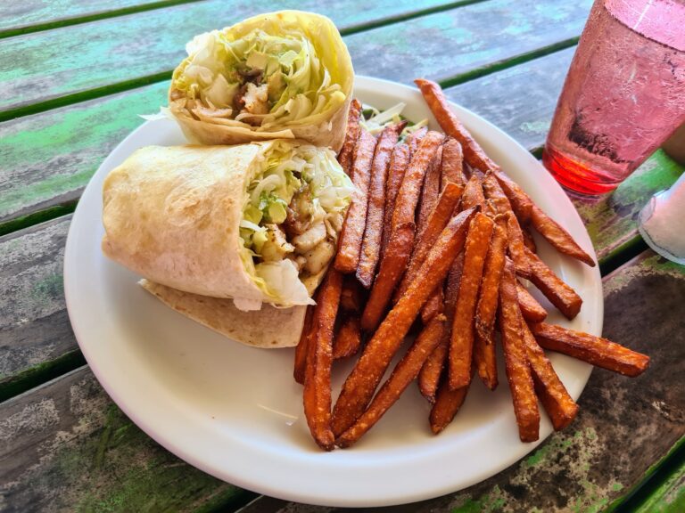 A plate of sweet potato fries, a wrap filled with lettuce and chicken, and a pink drink are displayed on a weathered wooden table at a restaurant in Roatan, Honduras.