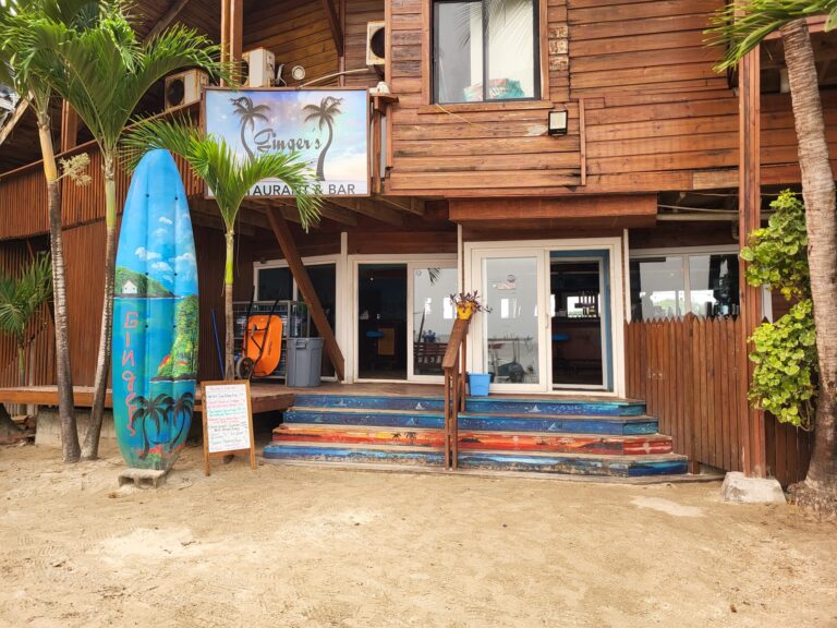 A wooden Caribbean-style building on Roatan, Honduras, features a sign for Ginger's Restaurant and Bar, with tropical plants, a colorful surfboard, and outdoor stairs leading to the entrance.