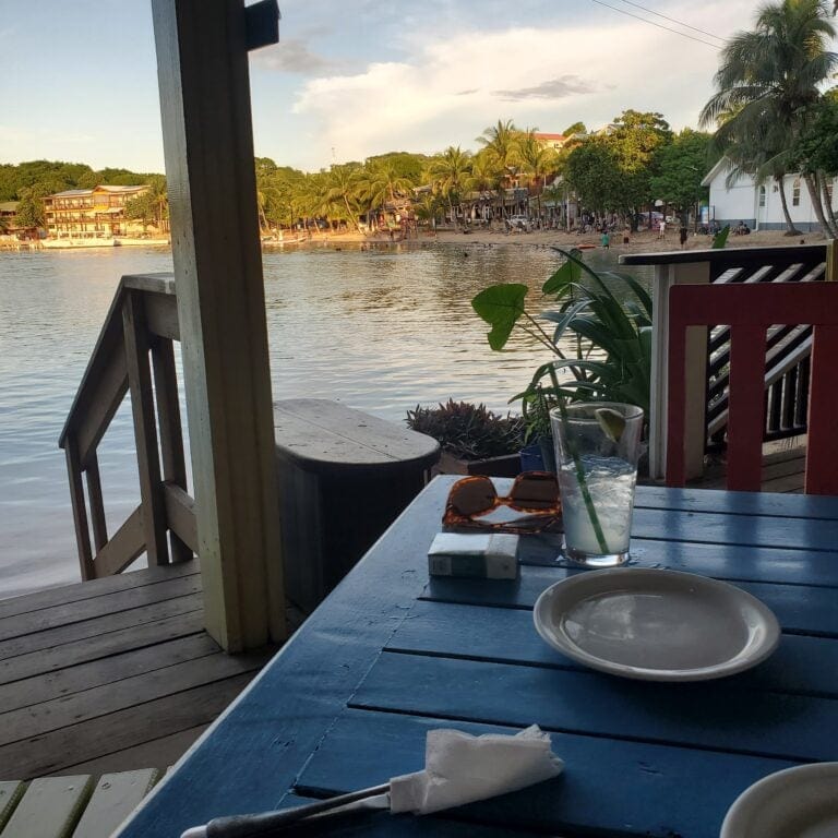 A view from a shaded waterfront deck in Roatan, Honduras, featuring a sand-covered beach with people, palm trees, and colorful buildings along the shoreline under a partly cloudy sky.