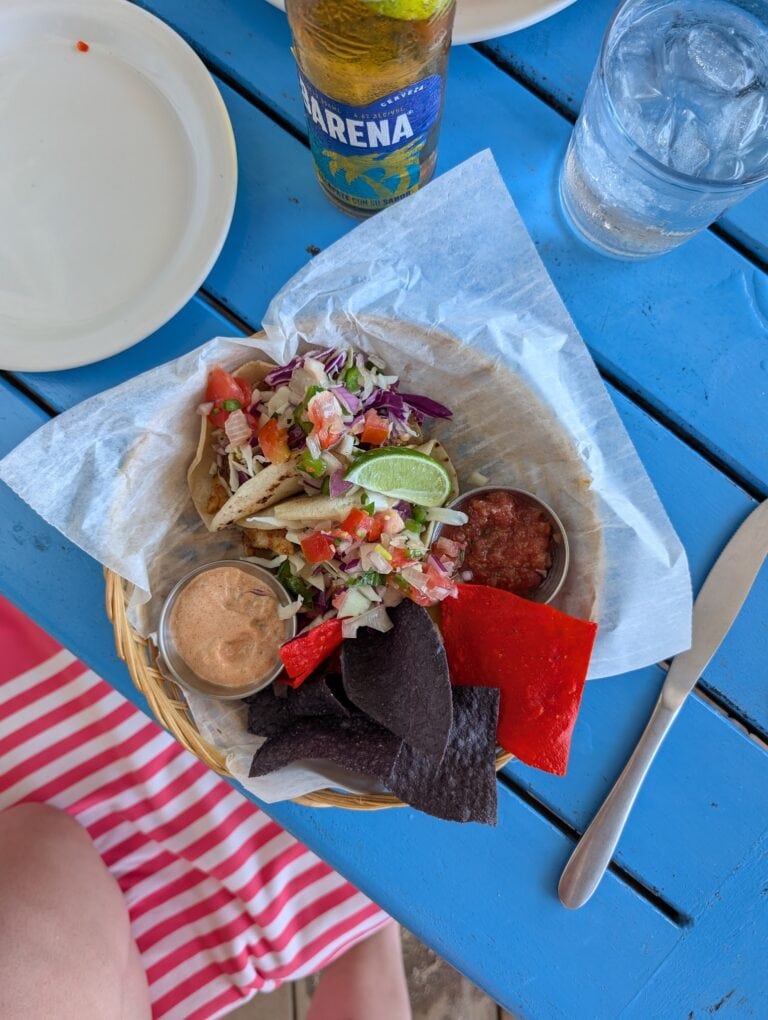 A basket of traditional Honduran food including tacos with fresh salsa and lime, crispy blue corn chips, and small bowls of sauce and refried beans, set on a bright blue table in Roatan, Honduras.