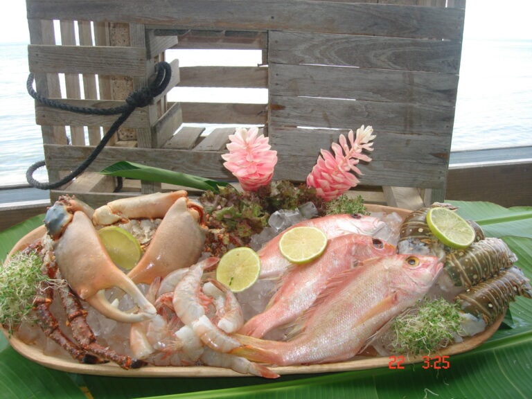 A seafood platter featuring lobster, shrimp, fish, and lime slices is displayed on a banana leaf in front of a weathered wooden structure with a rope handle, overlooking the water in Roatan, Honduras.