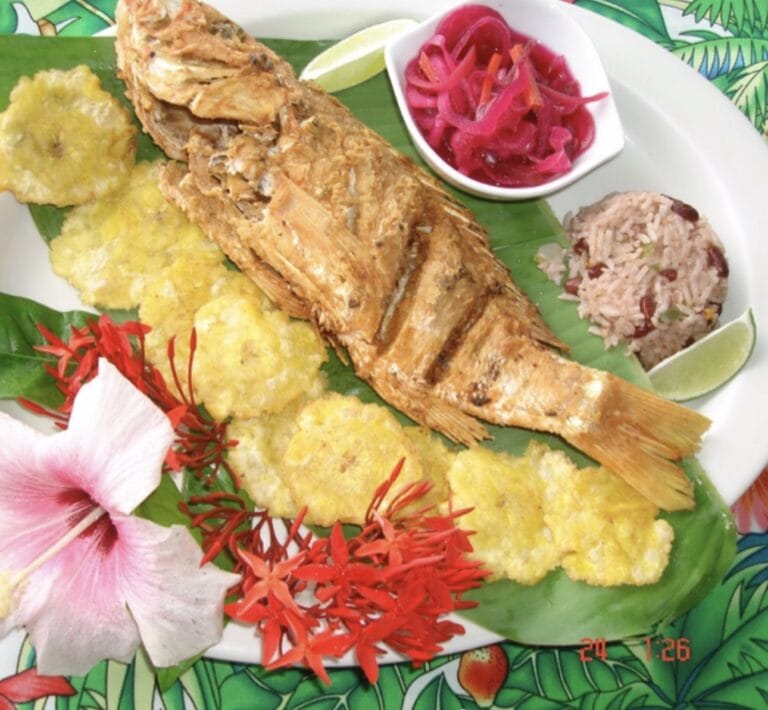 A plate of fried whole fish served with tostones, pickled red onions, a scoop of rice and beans, lime wedges, and decorated with tropical flowers on a green banana leaf in Roatan, Honduras.