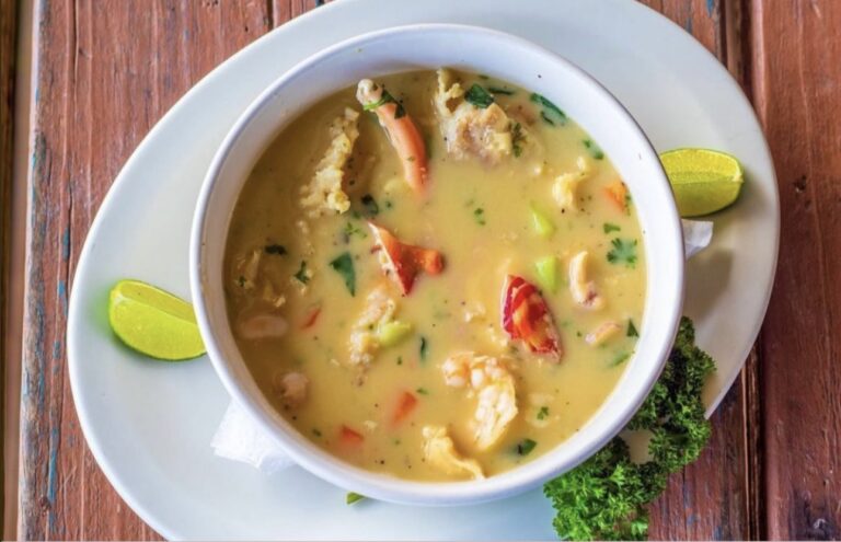A bowl of Honduran-style seafood soup with shrimp, vegetables, and herbs is placed on a white plate with lime wedges and fresh parsley on a wooden table in Roatan, Honduras.