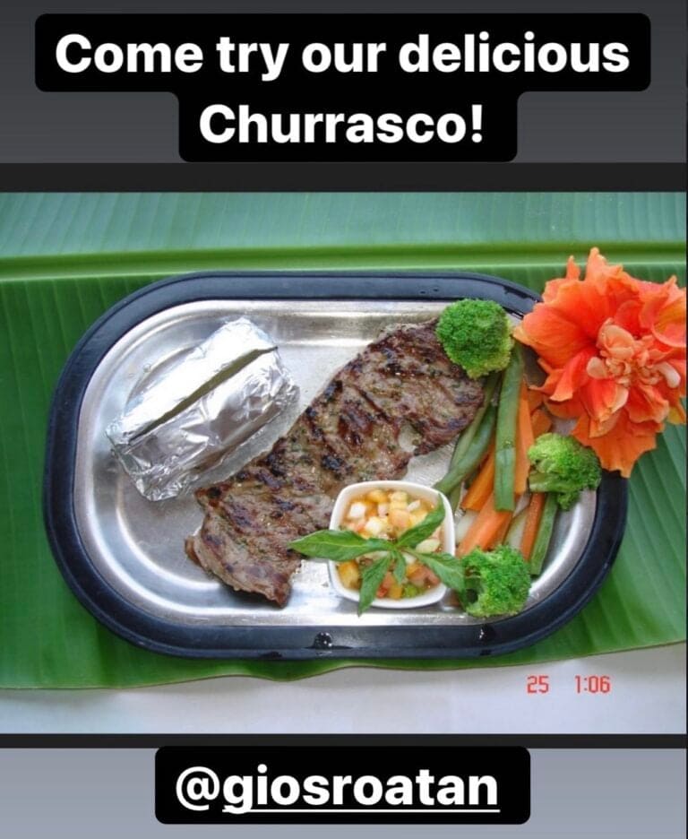 A grilled steak served on a stainless steel plate with a foil-wrapped baked potato, fresh vegetables, and a small bowl of salsa garnished with a basil leaf, all presented on a large green leaf, in a restaurant setting in Roatan, Honduras.