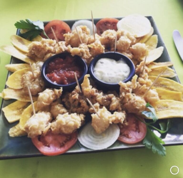 Fried chicken bites with sliced tomatoes, onions, banana chips, and dipping sauces served on a black tray at a restaurant in Roatan, Honduras.