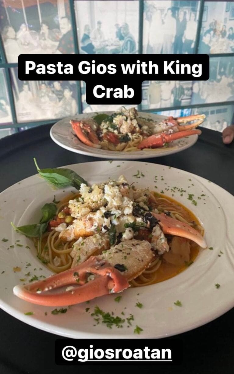 Two plates of pasta with king crab claws, garnished with herbs, are shown on a black tray and white plate, with a restaurant interior featuring windows and people in the background, located in Roatan, Honduras.