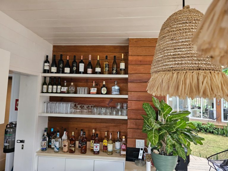 A liquor shelf with various bottles of wine, spirits, and glasses against a wooden wall inside a restaurant or bar in Roatan, Honduras, with a large plant and woven hanging lamp in the foreground.