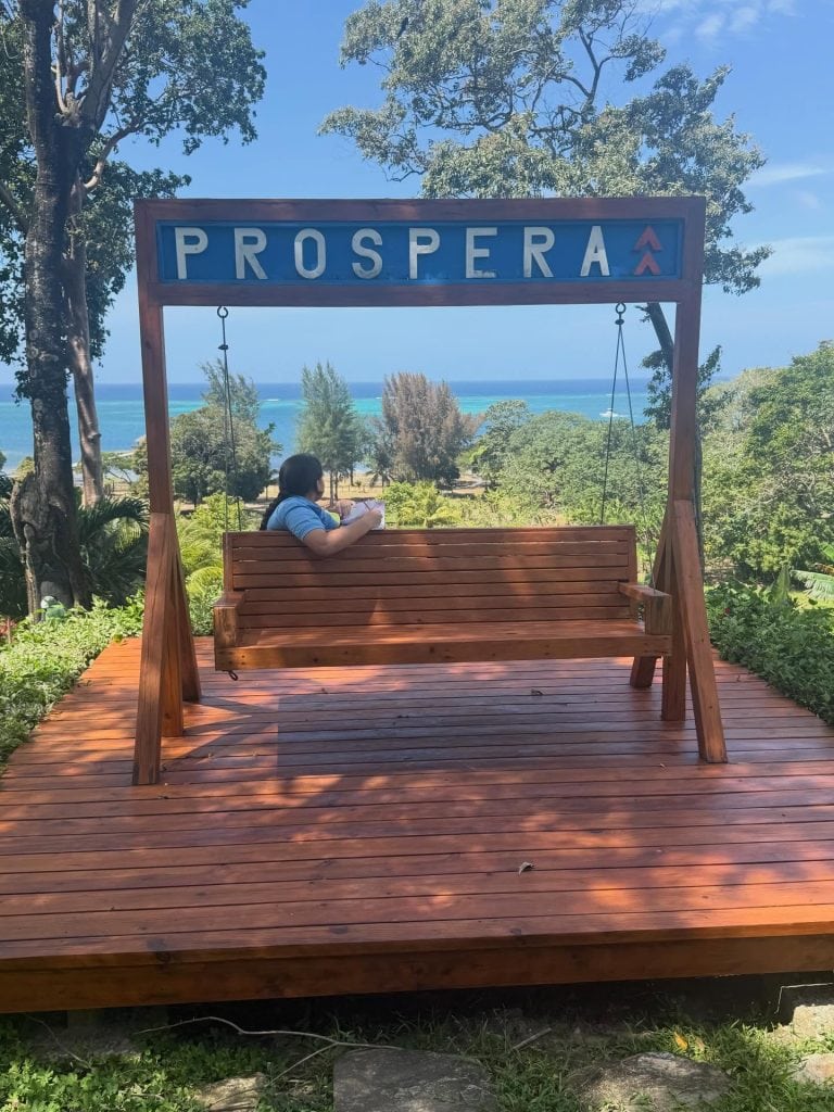 A woman sits on a wooden swing bench beneath a "Prospera" sign overlooking lush trees and the turquoise ocean near Roatan, Honduras.