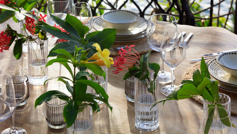 Elegant outdoor dining table on Roatan with flower arrangements in glass vases, surrounded by lush tropical greenery and decorated with plates, bowls, and glasses.