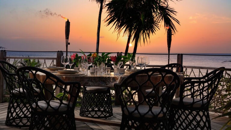 A luxurious outdoor dining setup on a wooden deck overlooking the ocean at sunset in Roatan, Honduras, featuring a round marble table, black wicker chairs, and a lush tropical backdrop with palm trees and tiki torches.