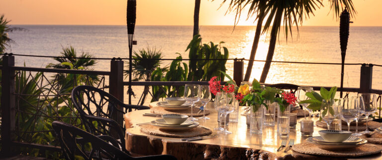 A beautifully set outdoor dining table overlooking the sunset on Roatan, Honduras with tropical plants and a view of the ocean in the background.